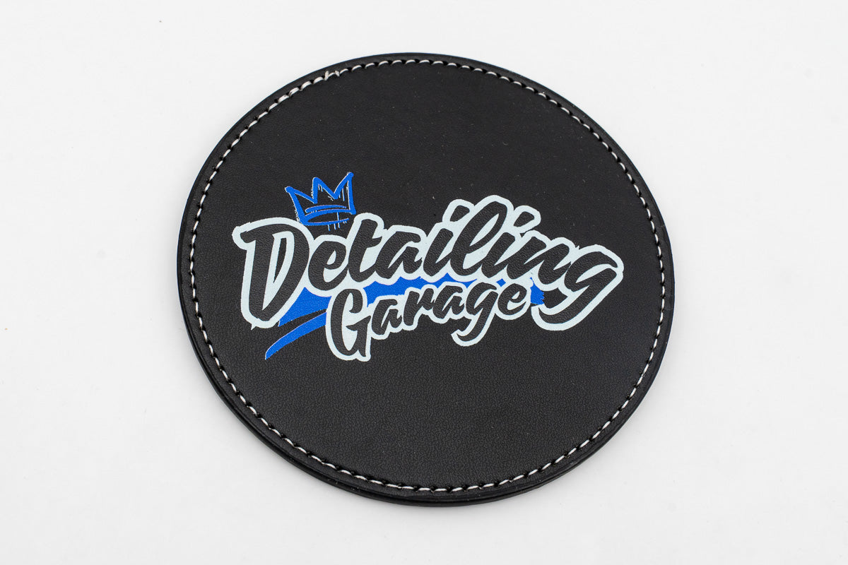 Round black coaster with a blue and yellow Detailing Garage logo.