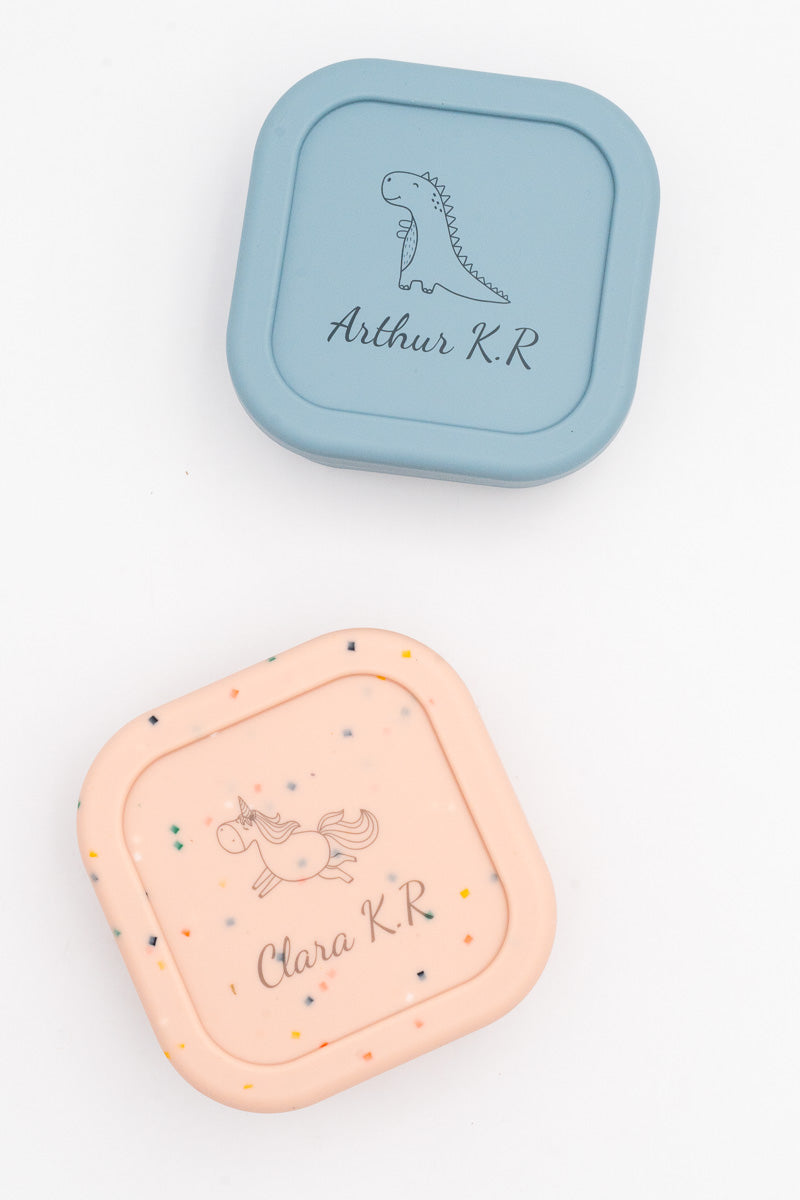 Two medium lunch boxes, blue and speckled pink, engraved with the names Arthur K.R and Clara K.R, a dinosaur and a unicorn