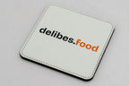Square white coaster with a black and orange delibes.Food logo.