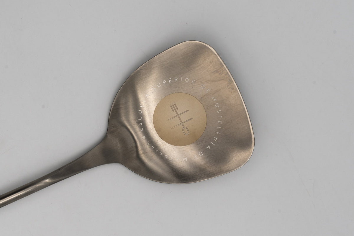 Stainless steel spatula with circular laser engraving of a hospitality school and dark handle
