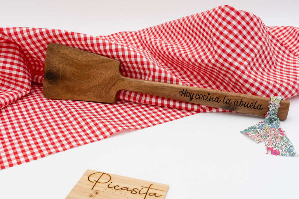 Espátula de madera oscura con mensaje Hoy cocina la abuela sobre mantel de cuadros rojos.