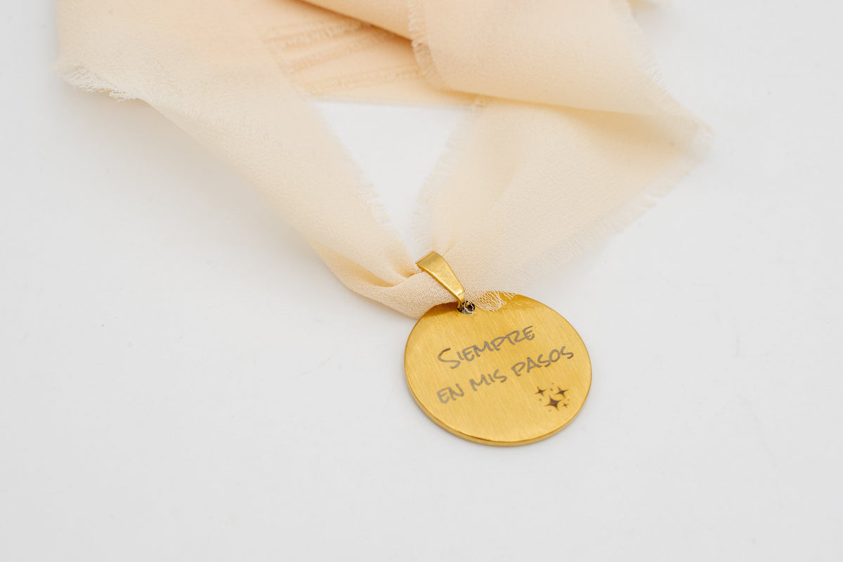 Gold charm with the phrase “Always in my steps” tied to a peach chiffon ribbon