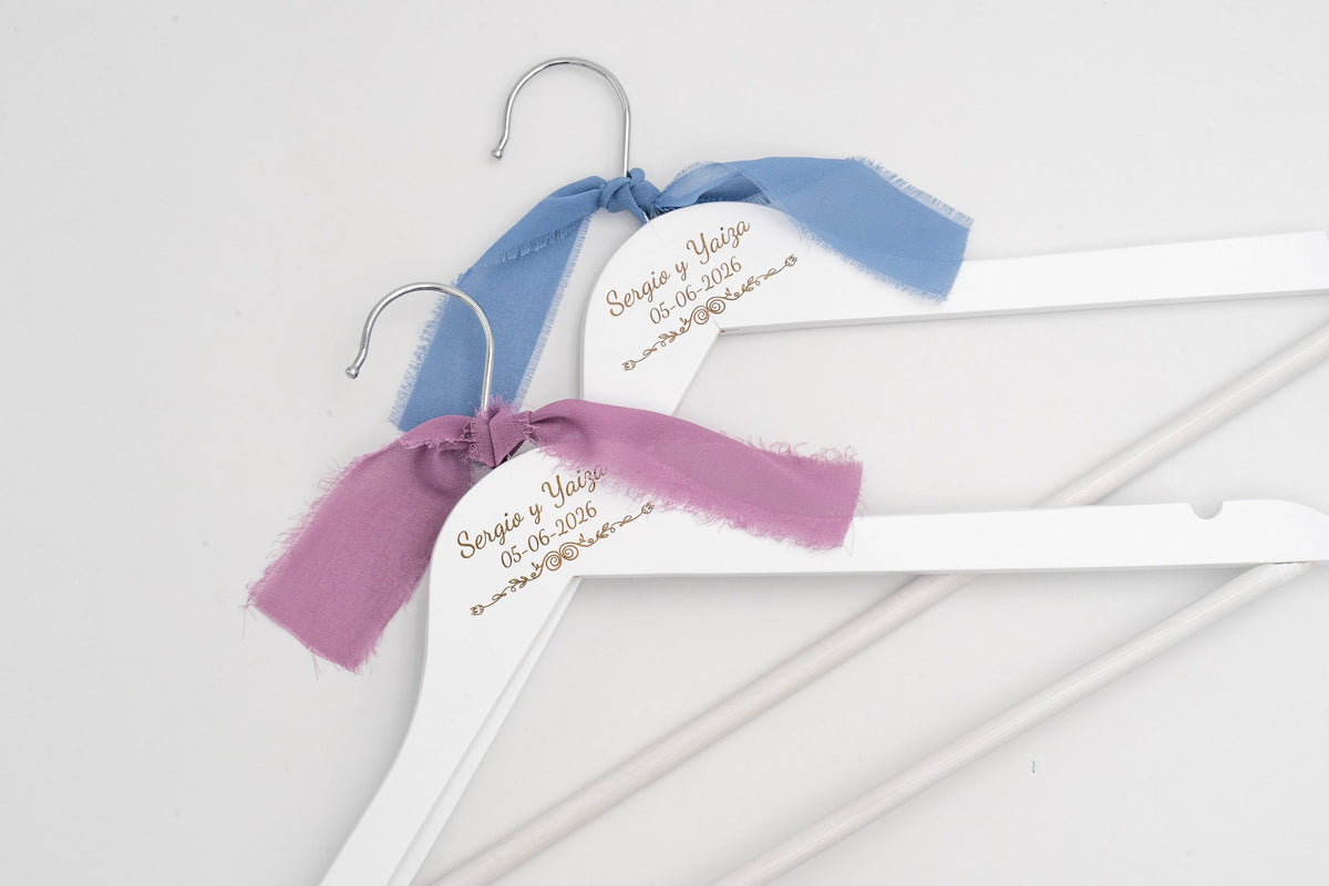 Two white wedding hangers with the couple's names engraved and blue and lilac chiffon bows